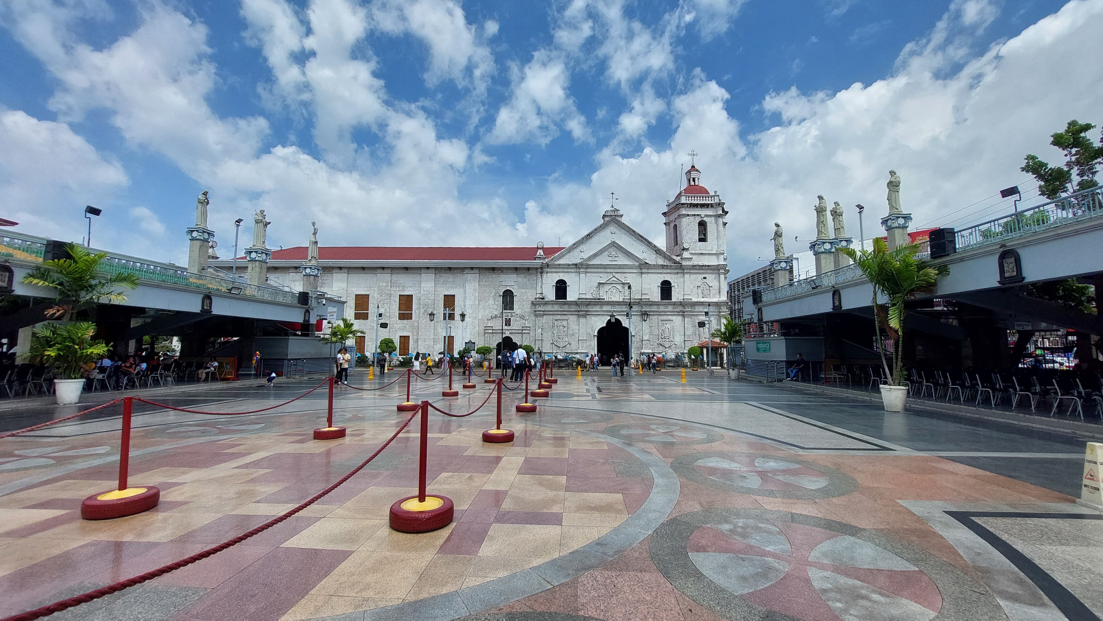 The basilica's belfry and facade seen across its wide pilgrim plaza, red rope stanchions standing on the patterned pavement