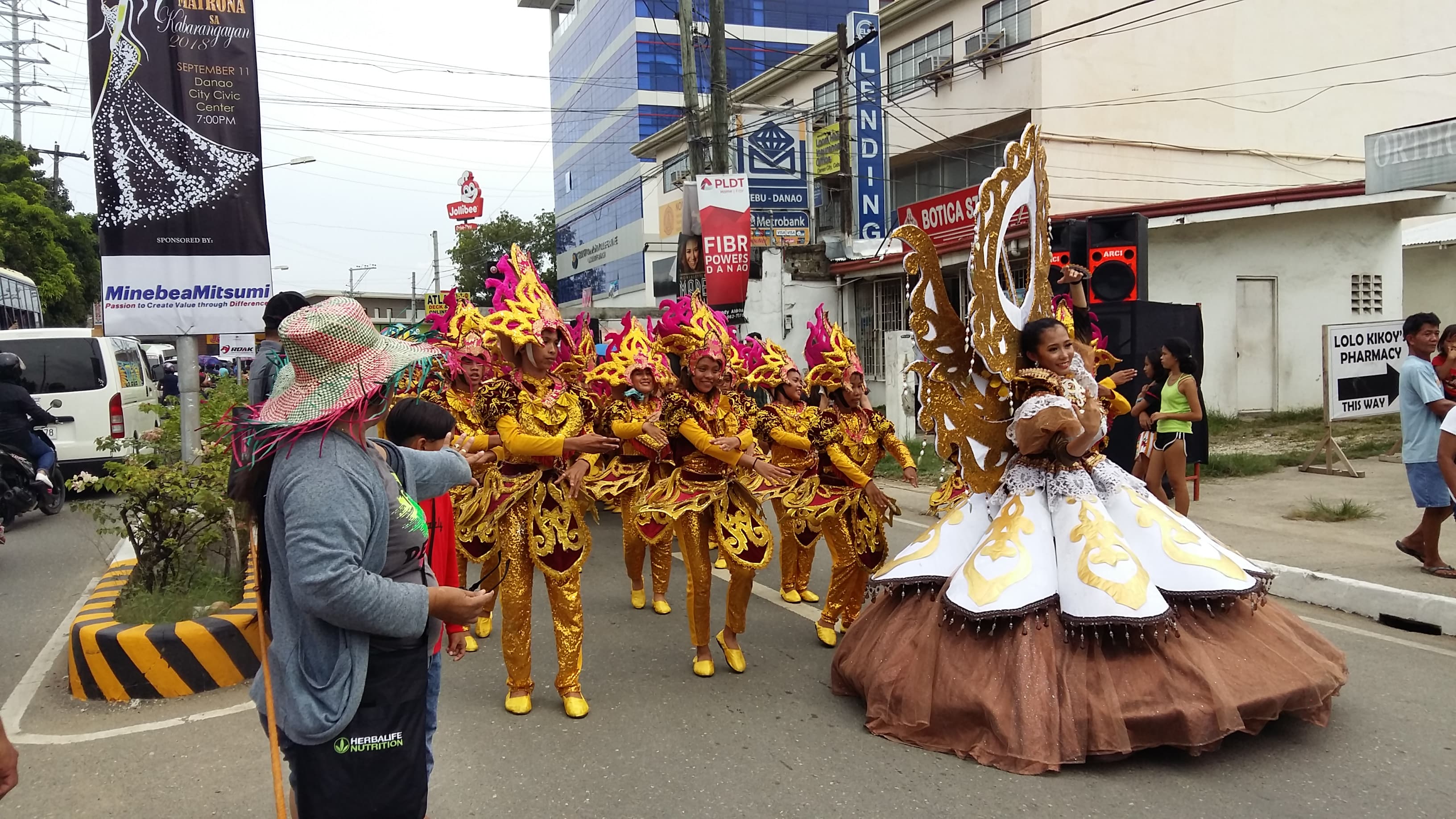 Karansa contingents in gold and pink costume dancing down a Danao City street, led by a performer in a wide brown and white gown