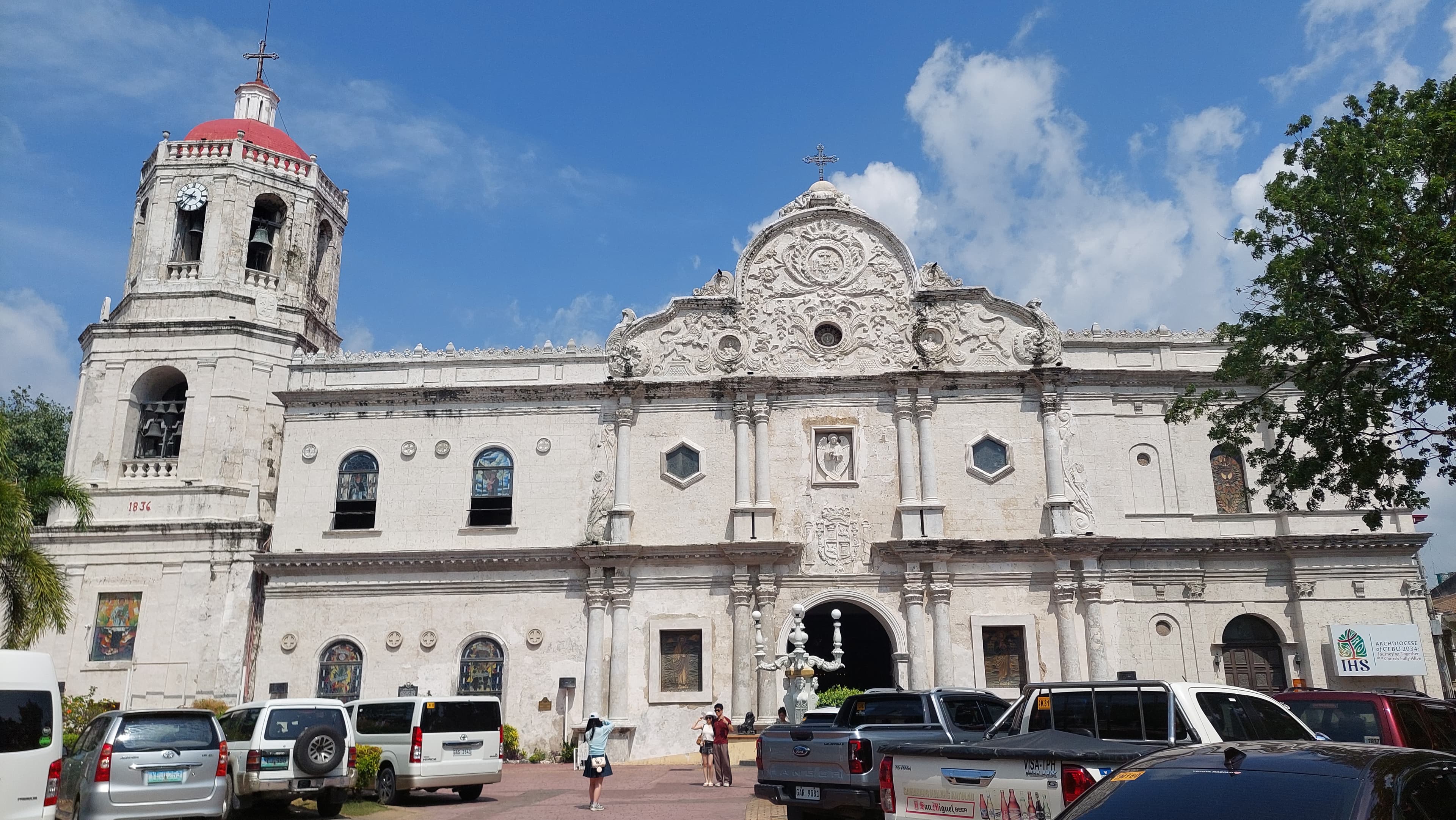 The cathedral's carved pale stone facade beside its separate red-domed belfry, seen over the parked cars on Independencia Street