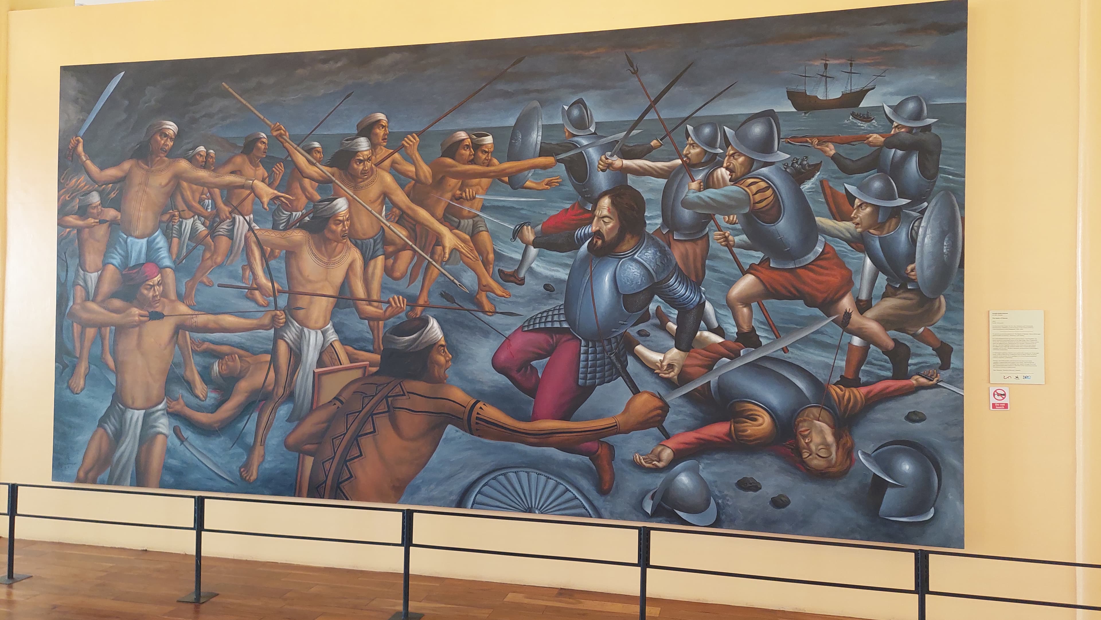 A large mural of the Battle of Mactan filling a gallery wall behind a low rail, showing Lapu-Lapu's warriors with spears and bows driving armoured Spanish soldiers back into the shallows, a galleon anchored offshore